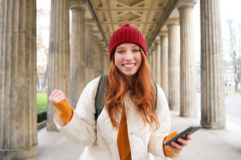 移动宽带人微笑红色头发的人女孩背包智能手机街持有移动电话应用程序