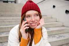 美丽的微笑红色头发的人女模型坐在街会谈移动电话智能手机应用程序调用在国外笑电话谈话