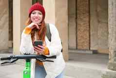 微笑年轻的红色头发的人女人移动电话租金e-scooter智能手机应用程序骑城市城市运输人概念