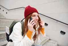 美丽的微笑红色头发的人女模型坐在街会谈移动电话智能手机应用程序调用在国外笑电话谈话