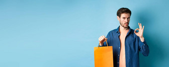 英俊的年轻的男人。推荐商店显示橙色购物袋标志站蓝色的背景