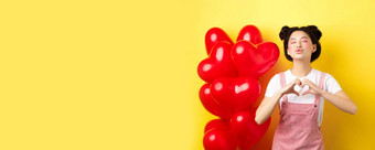 浪漫的女孩关闭<strong>眼睛皱纹</strong>嘴唇吻显示爱心手势站可爱的红色的气球接吻情人黄色的背景