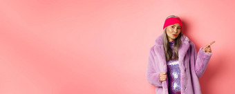 时尚的亚洲<strong>高级</strong>女人很酷的紫色的冬天外套指出手指显示广告时髦的相机粉红色的背景