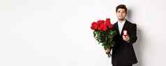 英俊的微笑男人。黑色的西装持有玫瑰订婚环使建议结婚站白色背景