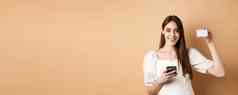 电子商务概念微笑女人显示塑料信贷卡购物在线移动电话站米色背景
