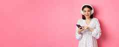 美丽的亚洲女人听音乐耳机移动电话微笑快乐相机站粉红色的背景