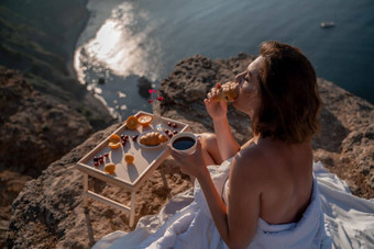 女人覆盖毯子床上放松看海景日出持有杯咖啡手前面表格水果羊角面包旅游热自由概念场景