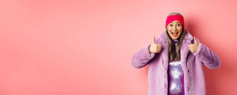 时尚购物概念快乐的亚洲女人显示竖起大拇指批准推荐促销活动站时尚的紫色的皮毛外套粉红色的背景