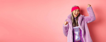 购物概念快乐的高级亚洲女人跳舞有趣的穿时尚的紫色的人造皮毛外套衣服赢得奖微笑站粉红色的背景