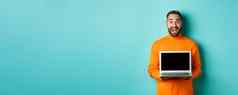 英俊的有胡子的男人。橙色毛衣显示移动PC屏幕展示促销站光蓝色的背景