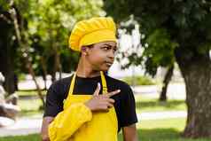 英俊的非洲少年烹饪点一边黑色的孩子烹饪厨师他黄色的围裙统一的微笑指出一边户外有创意的广告咖啡馆餐厅