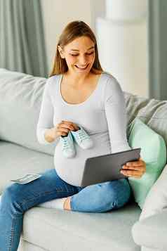 怀孕了女人妈妈。怀孕肚子鞋平板电脑沟通视频女人首页互联网视频卡尔在线年轻的持有显示小婴儿靴服装衣服