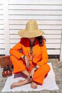 时尚的女人橙色西装他坐在地毯白色条纹背景手珠宝环手镯凉鞋袋站一边一边