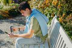 少年坐在板凳上秋天公园饮料咖啡热杯子电话肖像英俊的快乐的的家伙坐着板凳上新鲜的空气设备浏览媒体smm喝拿铁城市户外