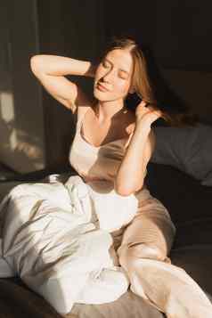 肖像女孩醒着的早....延伸床上太阳照窗口快乐年轻的女人问候阳光明媚的一天