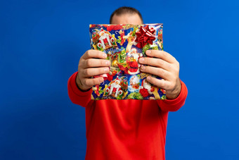 肖像快乐的男人。红色的毛衣持有礼物盒子前面房子孤立的蓝色的背景惊喜概念假期幸福情绪面部表达式庆祝活动