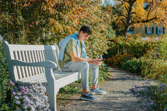 少年坐在板凳上秋天公园饮料咖啡热杯子电话肖像英俊的快乐的的家伙坐着板凳<strong>上新</strong>鲜的空气设备浏览媒体smm喝拿铁城市户外