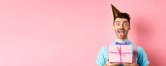 假期庆祝活动概念有趣的的家伙庆祝生日穿聚会，派对他持有生日礼物显示舌头快乐脸粉红色的背景