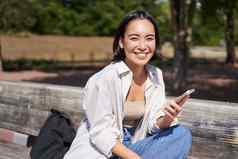肖像快乐年轻的亚洲女人会说话的移动电话公园坐着板凳上电话调用聊天活泼的