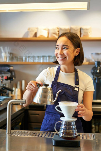 垂直拍摄微笑亚洲酒保咖啡师蓝色的围裙倒水小水壶酝酿咖啡计数器咖啡馆