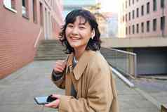 年轻的亚洲女人听音乐无线耳机智能手机街走城市阳光明媚的一天微笑幸福的
