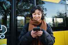 美丽的朝鲜文女孩学生公共汽车停止智能手机检查时间表阅读文本消息穿冬天衣服
