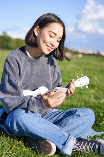 垂直拍摄微笑亚洲女孩智能<strong>手机玩</strong>尤克里里琴阅读和弦歌词唱歌放松在<strong>户外</strong>生活方式人概念