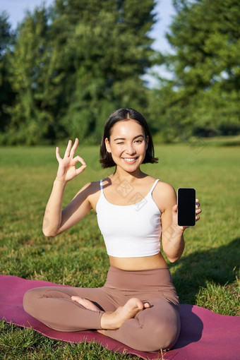 垂直拍摄亚洲女人显示标志智能手机屏幕<strong>推</strong>荐瑜伽<strong>培训</strong>在线冥想应用程序练习新鲜的空气公园