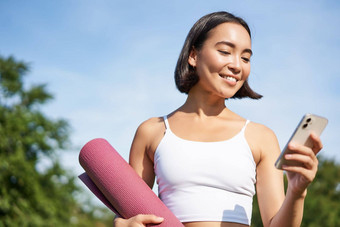 肖像微笑亚洲女人瑜伽席智能手机阅读应用程序站公园穿体育运动统一的