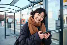 可爱的微笑亚洲女孩站公共汽车停止持有智能手机穿冬天夹克围巾女人通勤工作大学公共运输站路