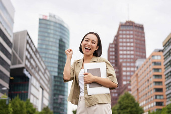 快乐的亚洲女孩城市中心显示<strong>万岁</strong>手势站移动PC庆祝