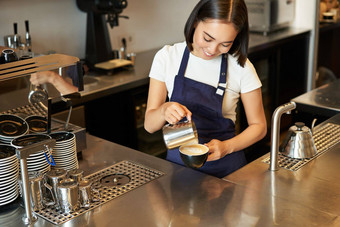 微笑亚洲女孩咖啡师咖啡馆工作人员倒蒸牛奶咖啡准备卡布奇诺咖啡拿铁艺术站蓝色的围裙计数器