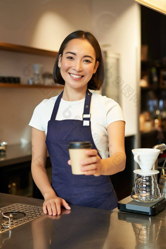 微笑亚洲女孩咖啡师给订单客户端持有外卖咖啡杯穿围裙工作计数器咖啡馆