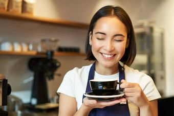 关闭微笑亚洲女孩咖啡师工人咖啡馆喝咖啡打破持有杯很高兴脸关闭眼睛享受喝