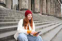 时尚的年轻的红色头发的人女人会说话的移动电话应用程序社会媒体应用程序在线智能手机坐在楼梯在户外