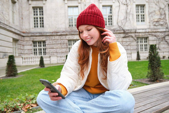 年轻的微笑红色头发的人女孩坐在板凳上智能手机应用程序读取新闻在线手表视频移动电话放松公园
