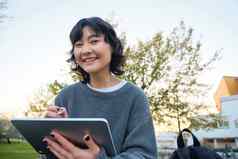 关闭肖像年轻的朝鲜文女孩坐在在户外公园持有数字平板电脑图形笔吸引了scatches灵感自然艺术微笑幸福的