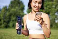 微笑健身女孩饮料水检查应用程序智能手机快乐保持水化新鲜的空气阳光明媚的一天公园