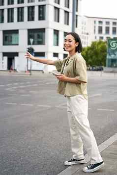 垂直拍摄年轻的女人抓出租车挥舞着司机路持有智能手机车分享应用程序站城市街