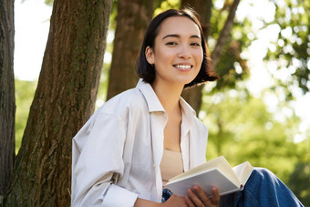 美丽的微笑女孩阅读书公园阳光明媚的一天坐着树草坪上放松在户外