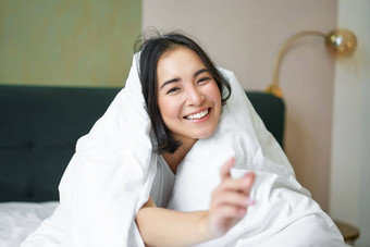 美丽的亚洲女人坐着床上覆盖白色<strong>羽绒被</strong>微笑享受快乐周末早....笑相机