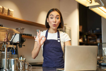 肖像咖啡师女孩亚洲女咖啡馆工人显示标志批准推荐smth快乐很高兴穿围裙站移动PC计数器