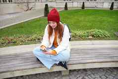 年轻的红色头发的人女孩学生坐在板凳上智能手机应用程序手表视频在线发送消息移动电话