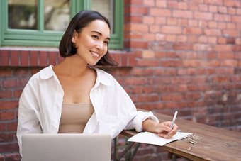 美丽的年轻的女模型坐着移动PC户外咖啡馆采取笔记写作<strong>日记工作</strong>项目家庭作业