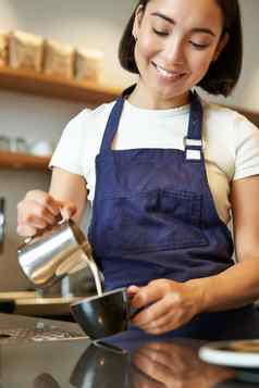 垂直拍摄浅黑肤色的女人亚洲女孩咖啡师穿围裙使咖啡准备订单咖啡馆倒牛奶杯卡布奇诺咖啡拿铁艺术