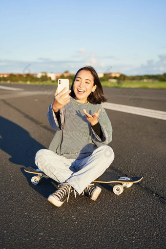 积极的青少年亚洲女孩需要自拍视频聊天移动电话应用程序坐在滑板记录视频博客