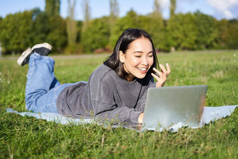 快乐亚洲女孩会说话的朋友移动PC视频聊天说谎公园毯子阳光明媚的温暖的一天