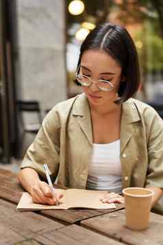 垂直拍摄年轻的亚洲学生女孩眼镜持有笔使笔记写作笔记本喝咖啡咖啡馆