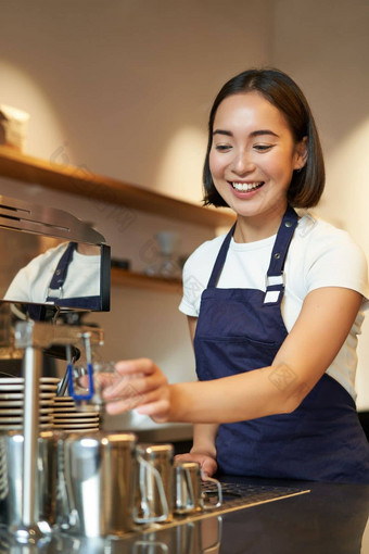 肖像可爱的咖啡师女孩工作计数器使咖啡热气腾腾的牛奶卡布奇诺咖啡穿咖啡馆统一的蓝色的围裙