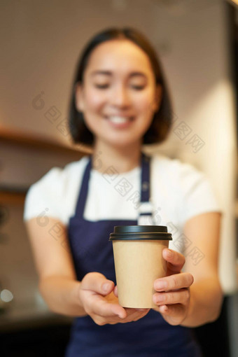 微笑亚洲女孩咖啡师给订单客户端持有外卖咖啡杯穿围裙工作计数器咖啡馆
